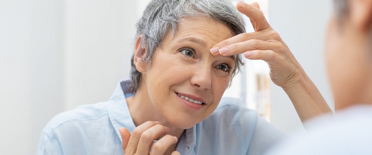 Mature woman looking her face and wrinkles in the bathroom mirror. Senior woman applying cosmetic lotion on skin between eyebrows while looking at mirror. Beautiful lady looking her face in the early morning.