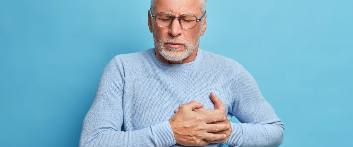 Senior man in spectacles presses hand to chest has heart attack suffers from unbearable pain closes eyes wears optical glasses poses against blue background. People age and problems with health